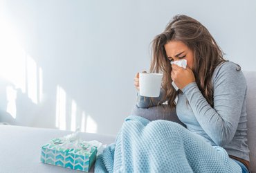 Frau auf dem Sofa, putzt sich die Nase und hat einen Becher in der Hand